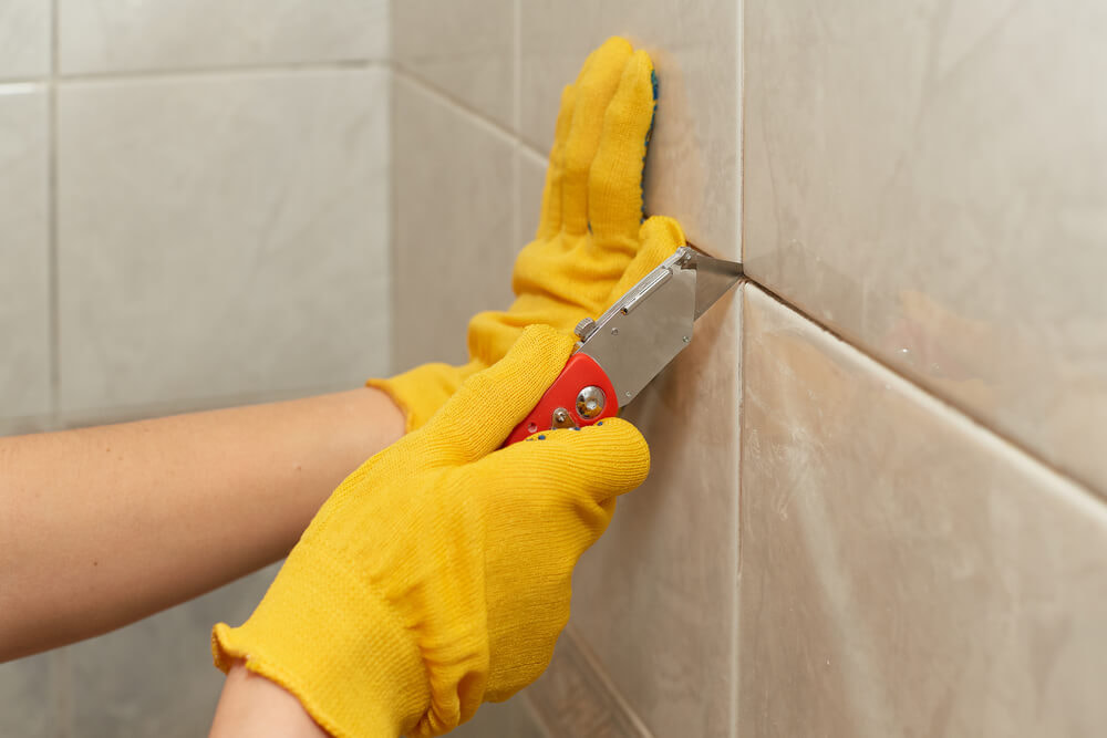 Gloved hands scraping old grout from wall tiles with a hand tool